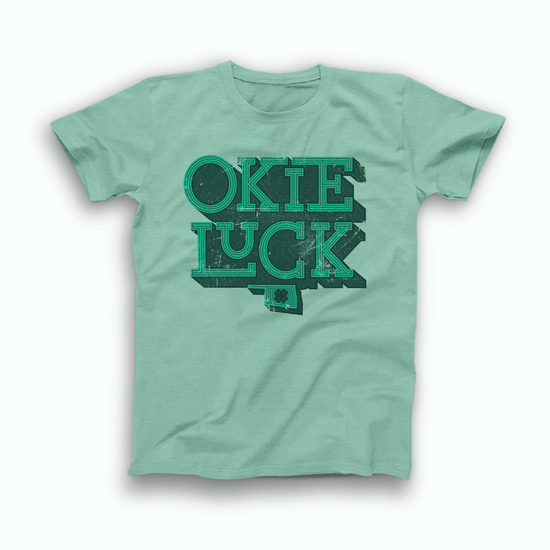 Mint green colored Oklahoma T-shirt. Screen printed in a distressed, vintage style in green with a dark green background is "OKIE" stacked atop "LUCK" with a small OK state symbol below and a 4 leaf clover inside the state.
