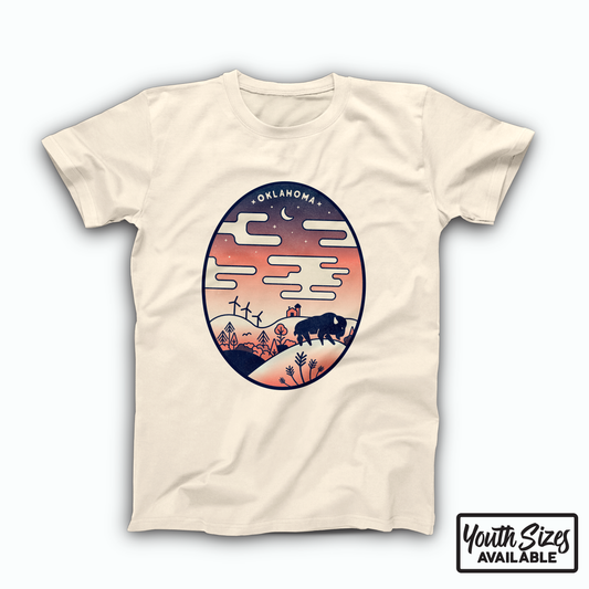 Natural colored Oklahoma t-shirt. Large, oval design across the front of the shirt in a indigo to peach gradient displaying an Oklahoma sunset over a hilly Oklahoma landscape, with clouds, stars, and the crescent moon above. The landscape includes wind turbines, a farm, plants and a bison.