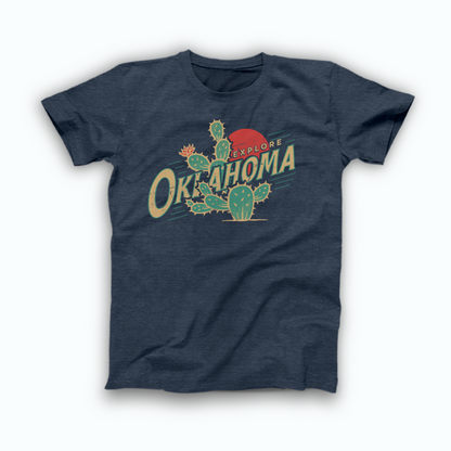 Heather Navy Oklahoma T-shirt. Design is of a large Prickly Pear Cactus with a red sun in the background and "Explore" in small font in front of the sun, beside the top of the cactus. And "Oklahoma" in large, bold font across the middle of the design at an upward angle, partially behind an arm of the cactus. Design in a distressed, vintage style.