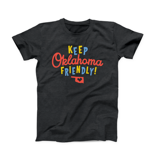 A dark heather grey color Oklahoma T-shirt. "KEEP" printed in alternating yellow and blue letters at the top. "Oklahoma" is red script font in the middle. "FRIENDLY!" at the bottom in alternating yellow and blue letters. The Oklahoma state shape with a heart cut out in the center is at the bottom of the design in red. 