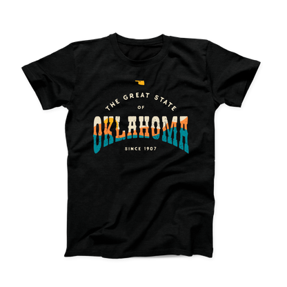 Black Oklahoma t-shirt. Small Oklahoma state shape at the top in yellow gold. "The Great State of Oklahoma since 1907" listed under the state. "Oklahoma" is the largest font on the shirt with a sunset over hills shown within the word in cream, yellow, orange, and teal. 
