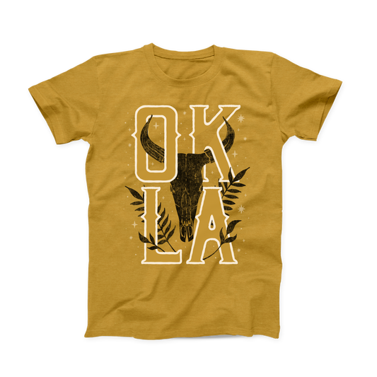 Gold Oklahoma T-shirt. Design is a distressed black Steer skull and leaf prints. In white is "OKLA" with little stars all around the design. OK is stacked above LA with the skull in the middle between the letters. 